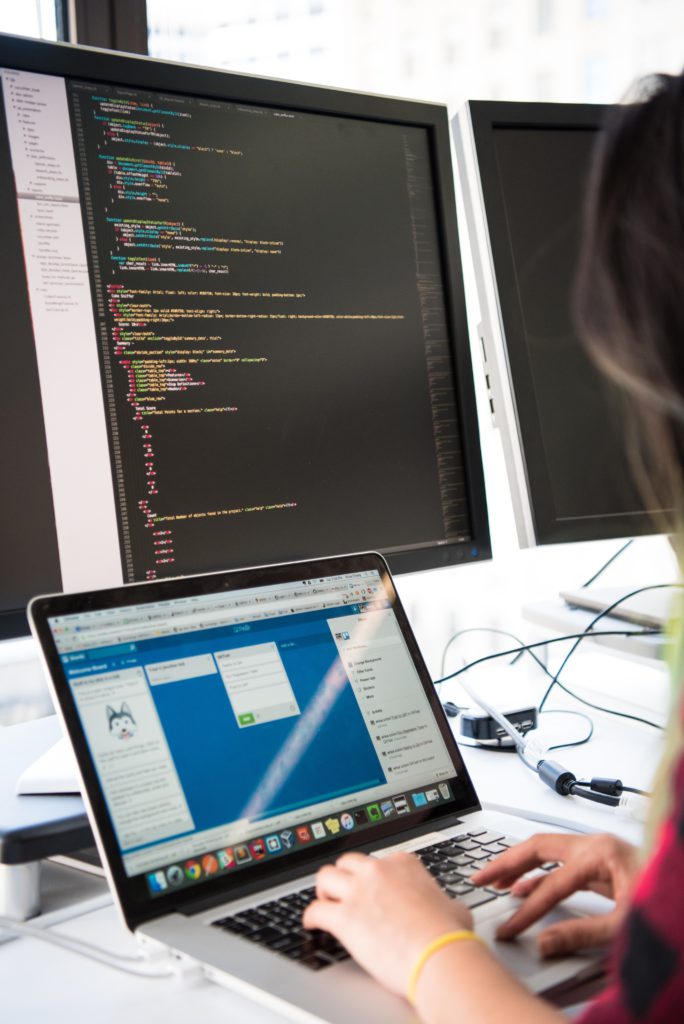 Professional IT services image showing Individual working on a laptop at a desk with two monitors displaying code in a bright, modern IT workspace. for managed IT services, cybersecurity, network infrastructure, cloud solutions, and business IT support.