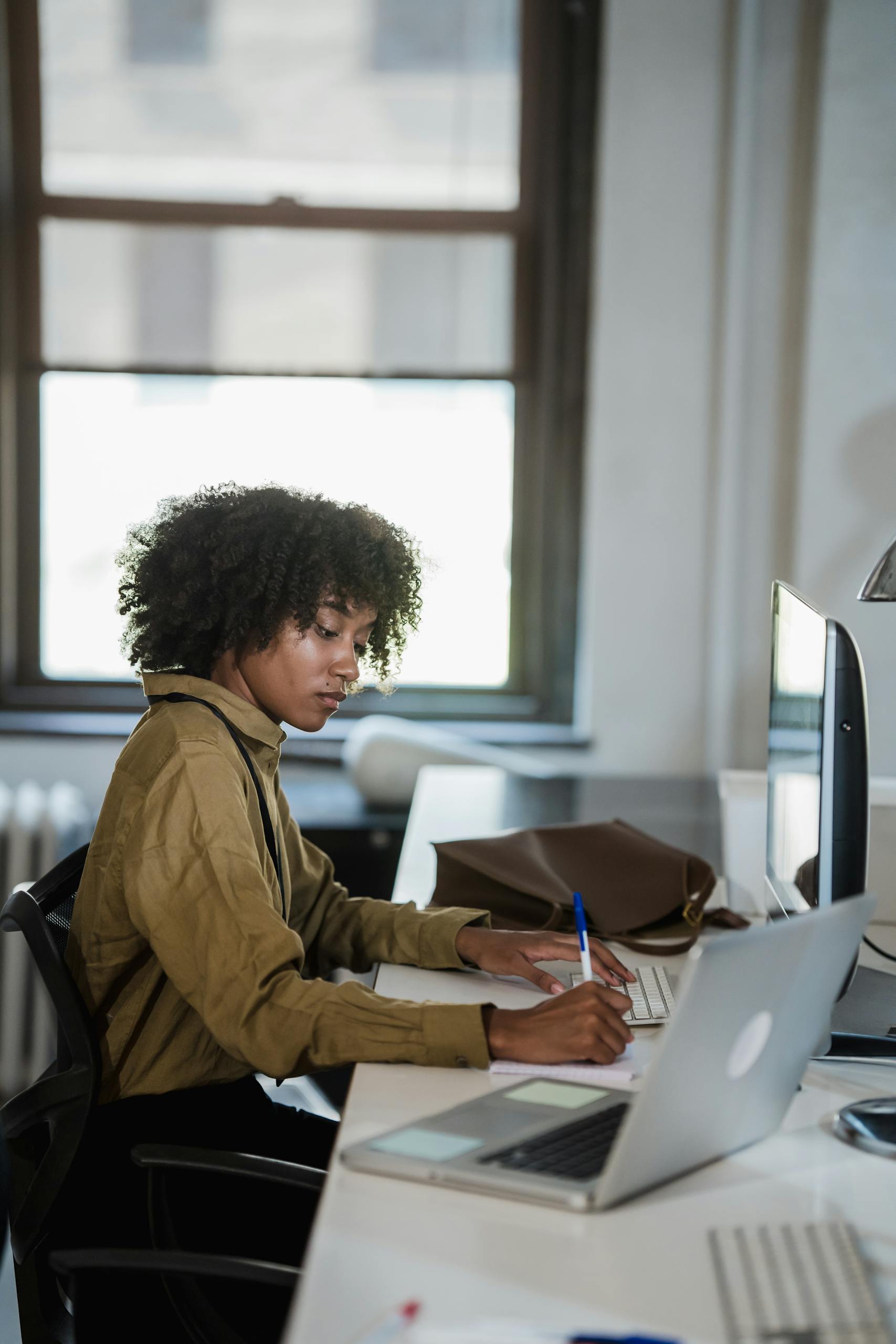 Young African American woman working at desk with laptop and monitor in office environment.